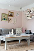 Hellgrau lackierter Couchtisch und Eckpolstersofa vor rosa getönten Wänden, oberhalb gerahmte Bilder
