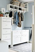 White narrow tallboy and chest of drawers under clothing hung from coat rack mounted on pastel blue wall