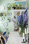 Candlesticks and blue flowers in glass jar on wooden table in front of display case and decorative wall plates on pale green wall