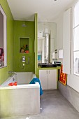 Badewanne, neben Trennwand eingebauter Waschtisch im Badezimmer mit Betonboden und teilweise grünen Wandfliesen