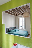 Blick durch Öffnung in grün gefliester Wand auf Doppelbett in schlichtem Schlafzimmer mit rustikaler Holzbalkendecke