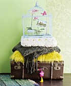 Stapel aus antikem Koffer, Kissen, Decken und nostalgischem Vogelkäfig vor pastellgrüner Wand