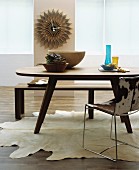 Wohnraum mit ovalem Holztisch, Sitzbank, Metallstuhl mit Tierfellbezug, Tierfellteppich und dekorativer Wanduhr