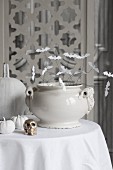 weiße Keramikschüssel dekoriert mit Papier Fledermäusen und kleiner Messing Totenschädel auf Tisch mit weisser Tischdecke