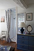 Table lamp and mirror on top of blue-painted chest of drawers next to curtain on doorway