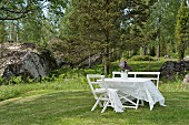 weiße Gartengarnitur mit Spitzentischdecke auf dem Rasen vor Wald und Felsen