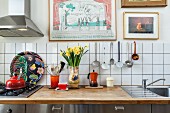 Vase of narcissus and kitchen utensils on worksurface against white-tiled splashback