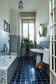 Blick durch offene Tür ins Bad auf dunkelblauen Fliesenboden und gemusterte Fliesen an Wand, im Hintergrund Zimmerpalme auf Schemel vor Fenster