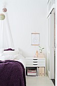 Modern bedside cabinet with drawers and purple blanket on bed below suspended globes