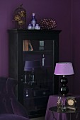 Table lamp with lilac lampshade in front of antique, glass-fronted cabinet against aubergine wall