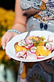 Frau trägt Teller mit Orangen-Radieschen-Salat