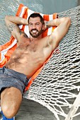 A laughing, topless man wearing shorts lying in a hammock