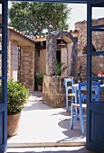 View through open terrace doors into sunny, Mediterranean-style courtyard with blue-painted chairs