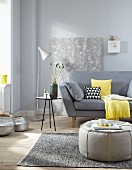 A pouffe in front of a sofa with cushion, a side table and a retro floor lamp against a wallpapered piece of chipboard