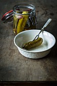 Pickled gherkins in a jar with one gherkin in a ceramic bowl on a wooden table