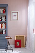 Picture frames on the floor against a wallpapered wall with a pin-stripped pattern in pink and white with a blue-grey shelf to the side