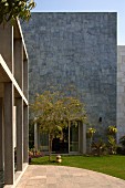 Tall, grey, stone façade with open terrace doors, garden and stone-paved area in front of concrete pillars