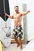 Lachender dunkelhaariger Mann in Badeshorts mit Surfbrett am Strand