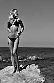 Blonde Frau in schwarzem Pailletten-Bikini steht auf Felsen am Meer (s/w-Foto)