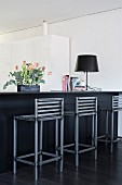 Flower arrangement and table lamp on counter with black-painted, metal barstools with backrests