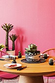 Runder gedeckter Holztisch vor pinkfarbener Wand mit Kakteen und Sukkulenten als Deko