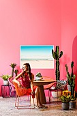 Retro holiday flair with lots of cacti: Young woman sits on Acapulco Chair at a round table in front of a pink wall