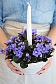 Purple African violet and white candle in ceramic bowl held in woman's hands