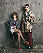 A brunette woman wearing a dark woollen coat, a skirt, blouse and fishnet tights and a man wearing a patterned coat and a suit