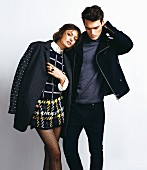 A brunette woman wearing a dark jumper with a glitter pattern, houndstooth patterned shorts and a short coat, and a man wearing a jacket, a turtleneck jumper and jeans