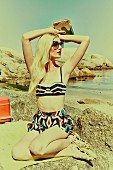 A woman wearing a bikini and geometric patterned shorts sitting on a beach