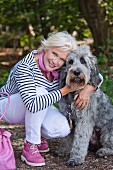 A blonde woman wearing a striped top, white trousers and pink shoes in a forest with a dog