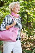 A blonde woman in the countryside wearing a striped top and white trousers holding a pink bag