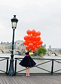Junge Fau in schwarzem Petticoat Kleid mit pinkfarbenem Gürtel und roten Luftballons auf einer Brücke