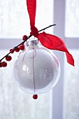 A white Christmas tree bauble decorated with a red ribbon and a sprig of holly berries