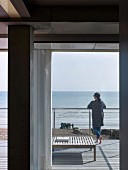View from house through open French window; woman leaning on terrace balustrade looking out over sea