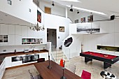 View across dining table and through fitted kitchen to pool table in open-plan interior with encircling gallery