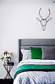 Bett mit grauem Kopfteil, Kissen und Bettwäsche in Grau und Grün, oberhalb an Wand Deko Tiertrophäe aus schwarzem Draht