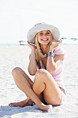 Junge blonde Frau mit Sommerhut, Schal und rosa T-Shirt sitzt im Sand am Strand