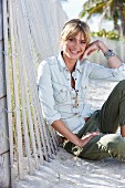 A young blonde woman sitting in the sand wearing a denim blouse and green trousers