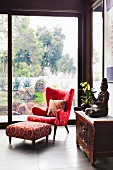 Elegant red reading armchair and footstool in front of floor-to-ceiling glazing with a garden view