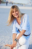 A young blonde woman on a beach wearing a light-blue shirt and white trousers