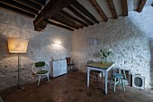 Kitchen table and wooden stools against white-painted stone wall below rustic wood-beamed ceiling