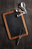 An old chalkboard, measuring spoons and a whisk on a dark brown wooden surface