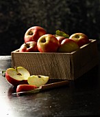 Rote Äpfel in Holzkiste, davor aufgeschnittener Apfel mit Messer