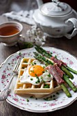 A savoury waffle with a fried egg, asparagus wrapped in ham and chives