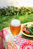 A glass of Pils on a garden table next to a plate of summer salad
