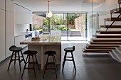 White, designer kitchen with island counter, staircase and glass wall with garden view