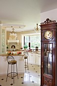 Antique, elegant long-case clock and open-plan, white, country-house kitchen with chandeliers