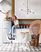 Young woman sitting on kitchen counter, tulip table with matching chairs and peacock throne, above pendant lamp with glass shades