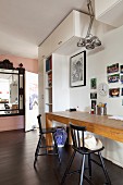 Narrow table made of pale wood and black bar stools below collection of photos on wall